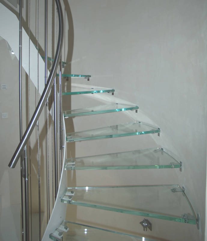 Escalier à limon métal cintré avec marches en verre clair. Rampe composée de poteaux et main courante en inox Ø 42 mm, les câbles inox verticaux sont tendus entre le sol et la main courante.
