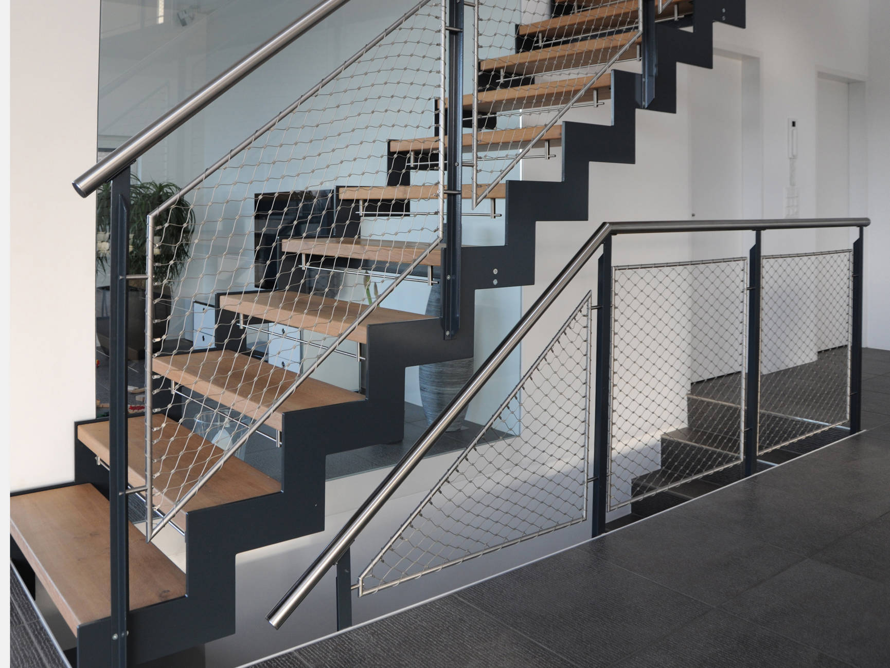 Metallwangentreppe abgetreppt mit Massivholzstufen in Eiche.  Geländer mit T-Pfosten und CNS-Handlauf. Geländerfüllung als Netz mit Inox-Seilen, das sogenannte Webnet. Kinderschutzleisten aus Edelstahl.