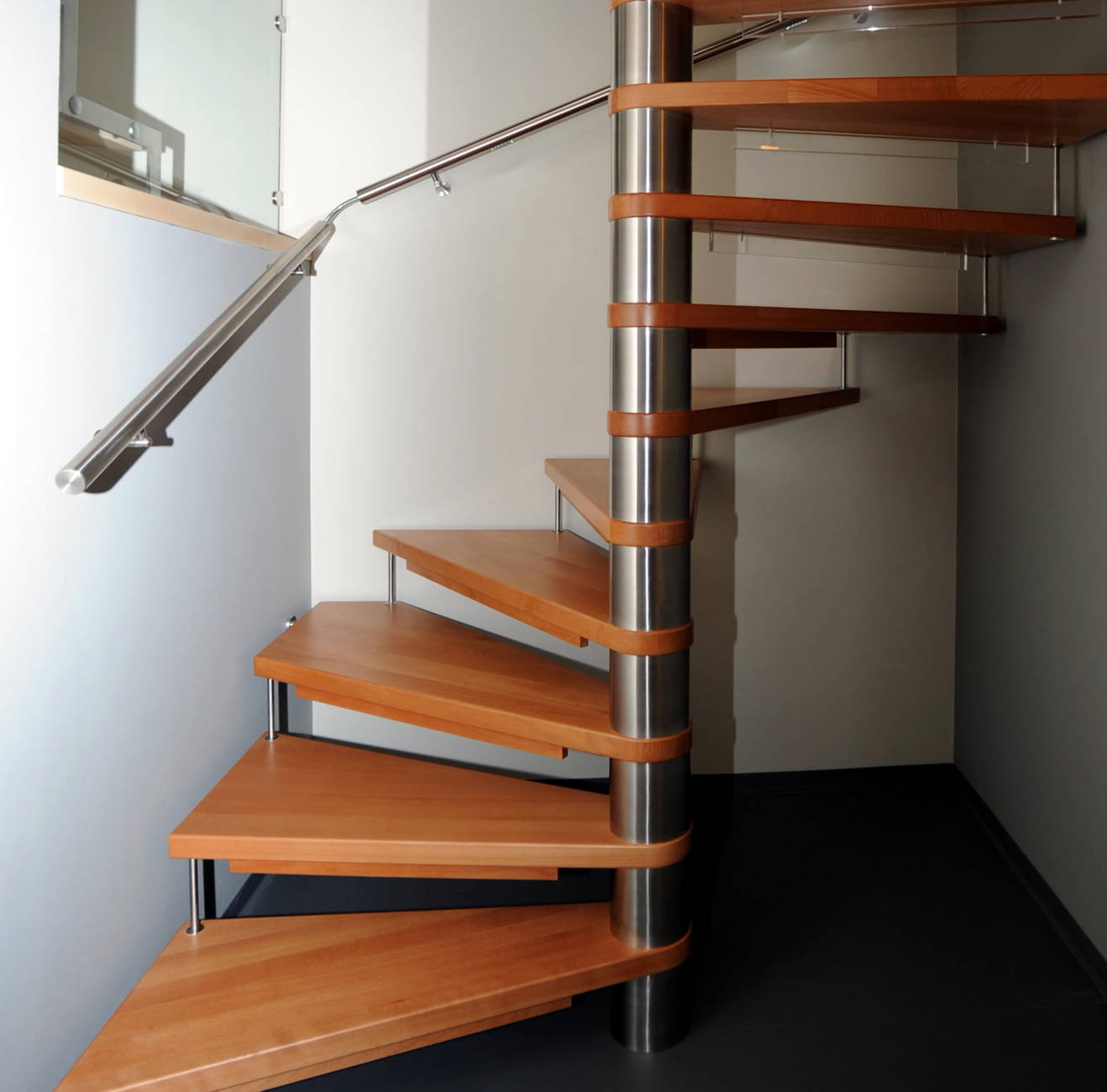 Spindeltreppe in Buche gedämpf, farblos lackiert. Säule und Stufenverbinder in Edelstahl. Kinderschutzleisen in Holz und Plexiglas. CNS-Handlauf an Wand.