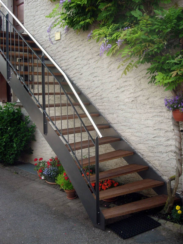 Escalier d'extérieur avec limons métalliques et marches en bois. Balustrades montantes avec poteaux en fer T, lisses hautes et basses, balustres verticaux en acier laqué, main courante en inox.