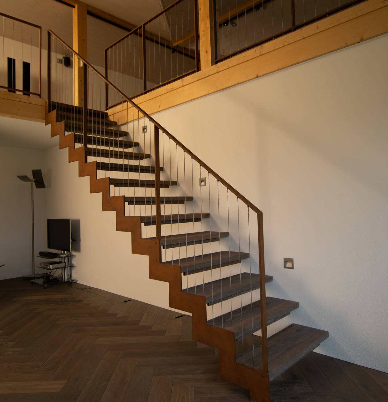 Metallwange unten und oben abgetreppt und Geländerrahmen in Rostoptik, vertikale Inox-Seile und Massivholzstufen.