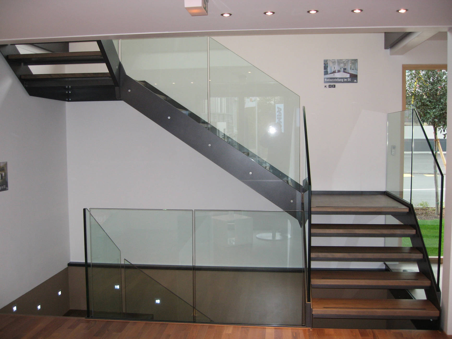Metallwangentreppe mit Massivholzstufen, jeweils einem Gummigleitschutzstreifen auf der Stufenvorderkante und einem Brandschutzblech unter der Stufe. Glasbrüstungen zwischen 2 Metallwangen montiert (Metallsandwichkonstruktion). 