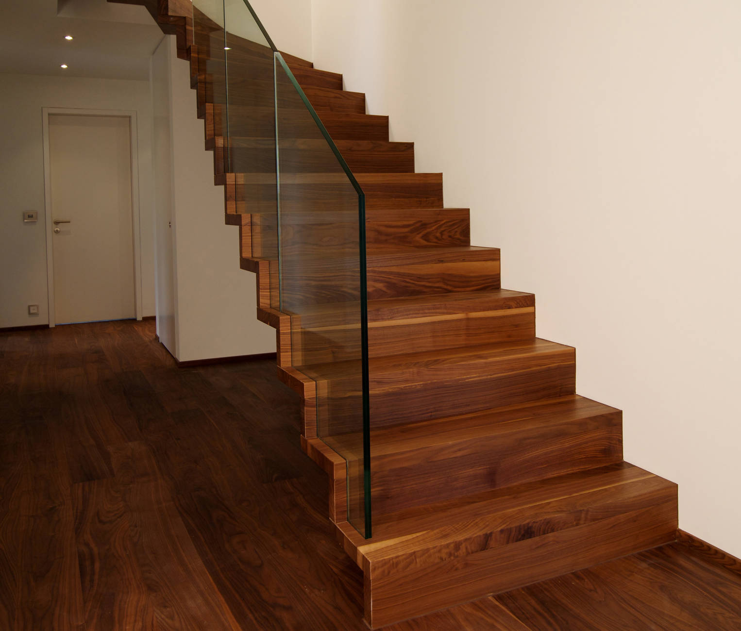 Faltwerktreppe aus amerikanischem Nussbaum natur farblos lackiert, mit der Wand verschraubt. Glasbrüstung lichtseitig ins Massivholz eingenutet.