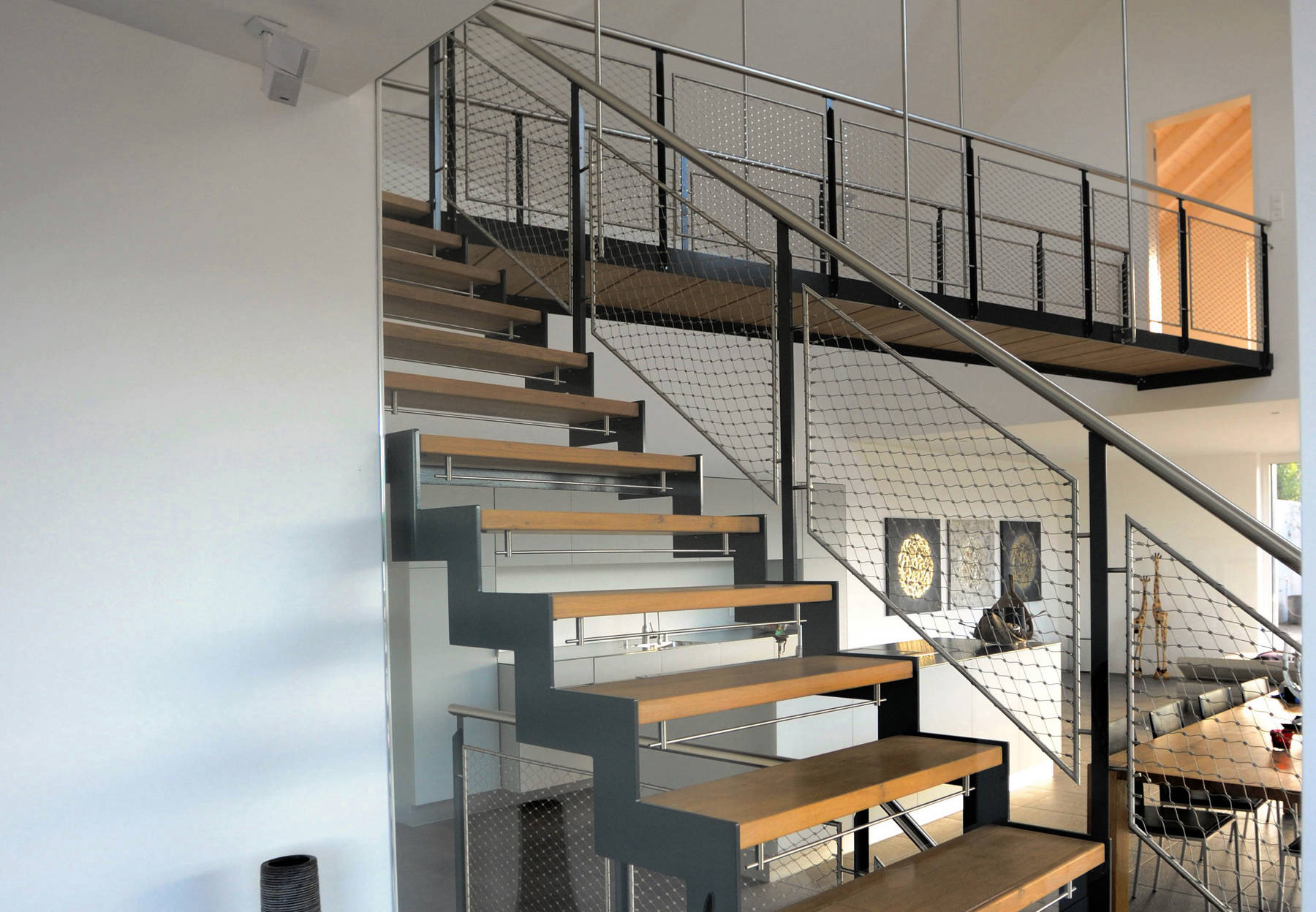 Metallwangentreppe abgetreppt mit Massivholzstufen in Eiche.  Geländer mit T-Pfosten und CNS-Handlauf. Geländerfüllung als Netz mit Inox-Seilen, das sogenannte Webnet. Kinderschutzleisten aus Edelstahl.
