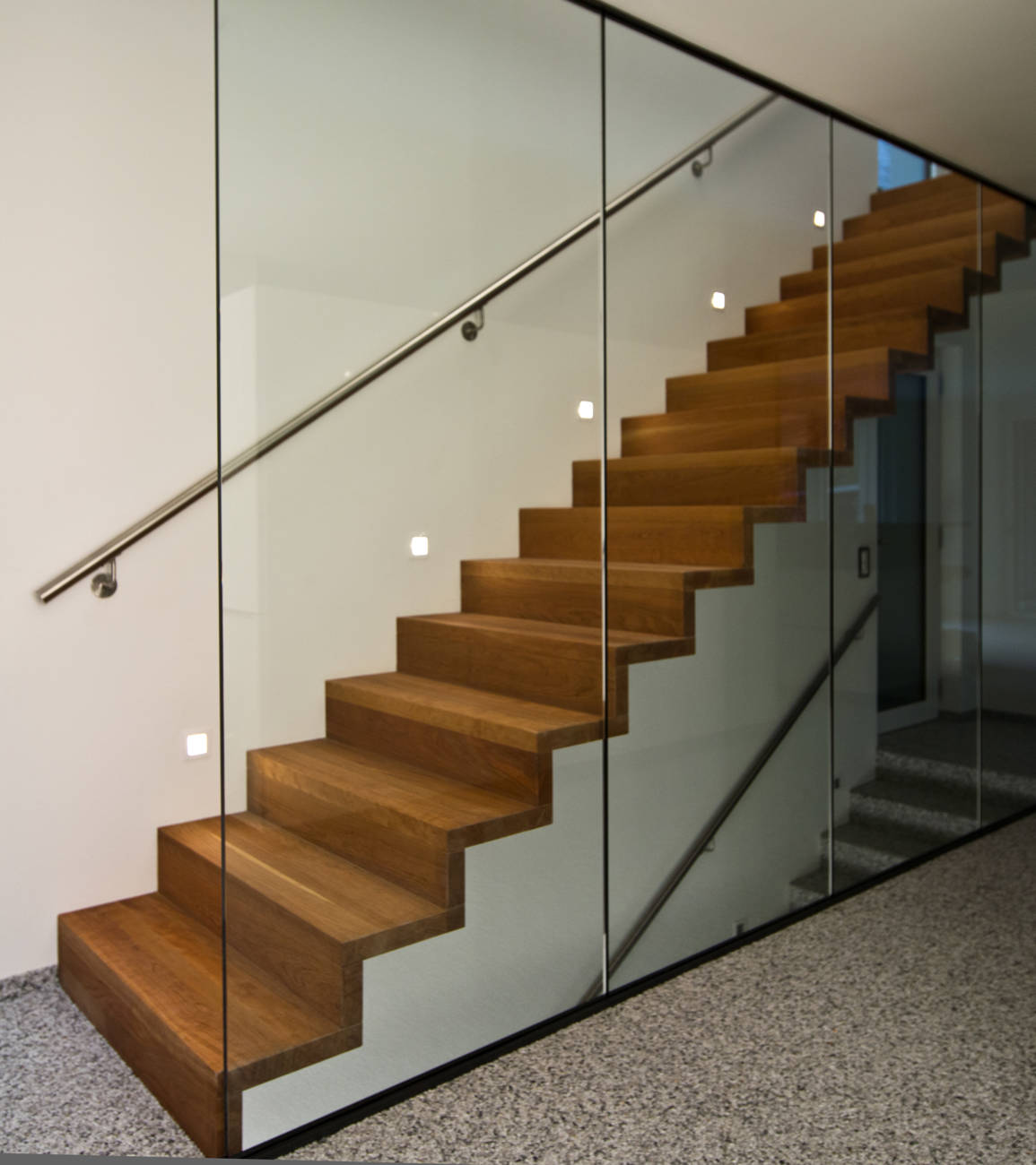 Faltwerktreppe in Amerikanischem Kirschbaum geölt, mit der Wand verschraubt. Glaswand von Boden bis Unterkant Decke in Metallkonstruktionen montiert. CNS-Handlauf an Wand.