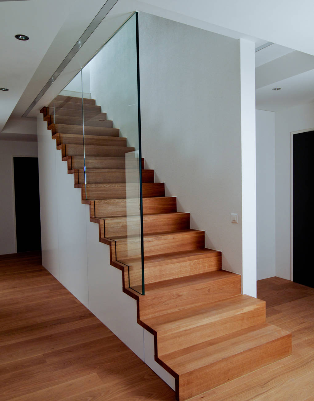 Stufen und Futterbretter in Eiche natur geölt als Faltwerk ausgebildet und auf zwei Holzsattel befestigt. Glas unten in Stufen und Futterbretter eingenutet und oben in Metallkonstruktion geklemmt. Die Wand hat keine tragende Funktion.