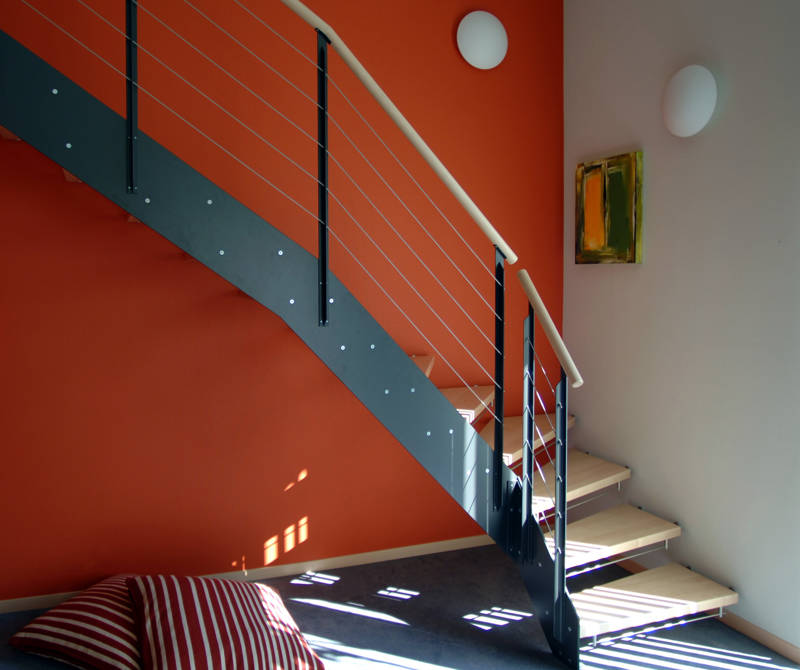 Loft-Treppe, Buchestufen innen mit HPL-Vollkernplatte, aussen direkt in Wand befestigt. Geländer mit T-Pfosten und Inox-Seilen und Holzhandlauf. Kinderschutzleisten in Edelstahl.