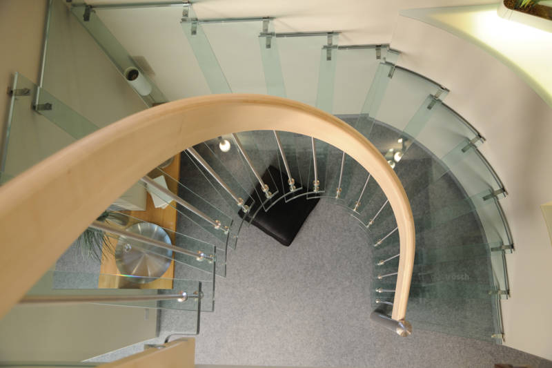 Escalier autoportant courbe avec marches en verre (VSG 31mm), rampe avec main courante courbe en érable lamellé collé, balustres et poteau de départ en inox. 