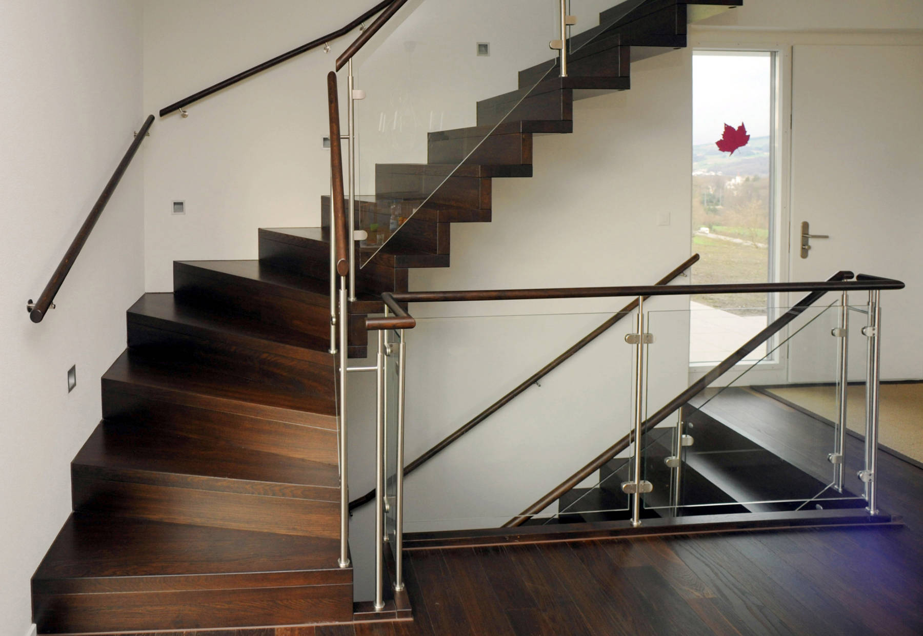 Faltwerktreppe aus Eiche gebeizt, mit der Wand verschraubt. Geländer mit Edelstahlpfosten, Holzhandlauf und Glasfüllungen. Holzhandlauf an Wand.